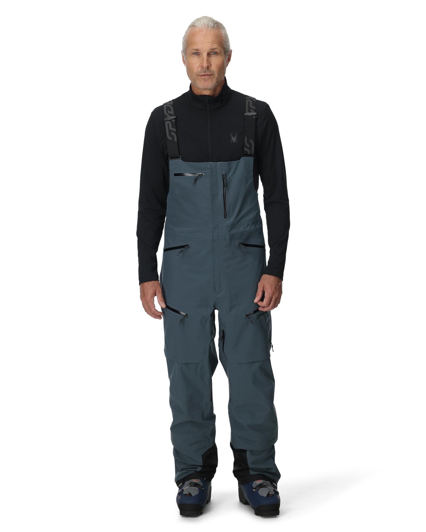 Man wearing a black long-sleeve shirt and blue bib overalls on a white background