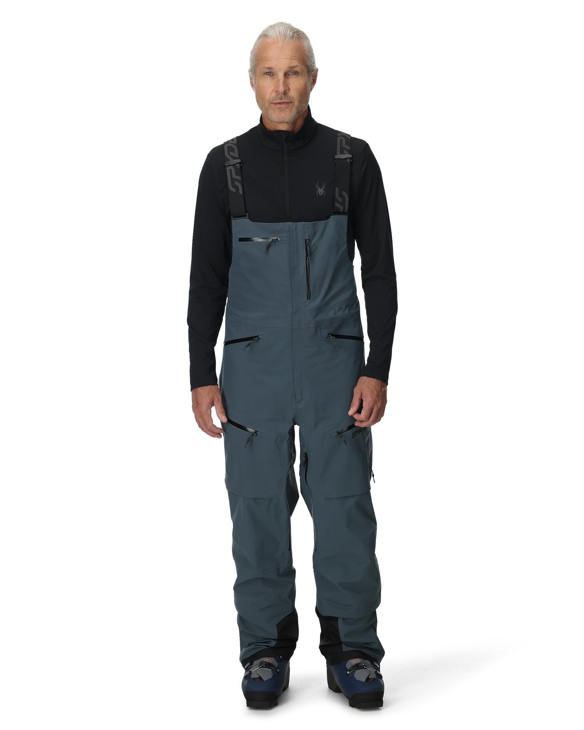 Man wearing a black long-sleeve shirt and blue bib overalls on a white background